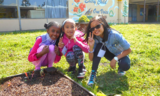 Three girls together in the grass smiling, one Black, one Latina, one Asian American